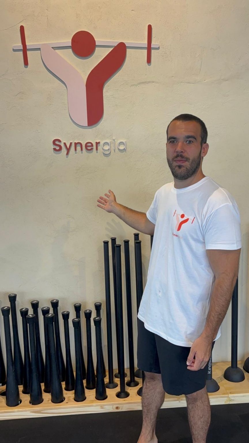 Rubén junto al logo de synergia. Se situa justo delante del logo vestido con una camiseta blanca con el logo del centro.